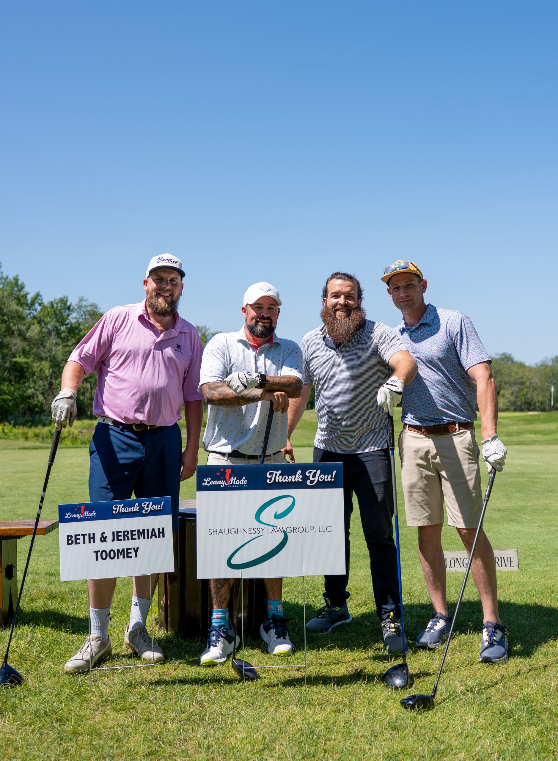 Foursome of four men posing at the long-drive tee behind the Beth and Jeremiah Toomey and Shaughnessy Law Group sponsor signs.