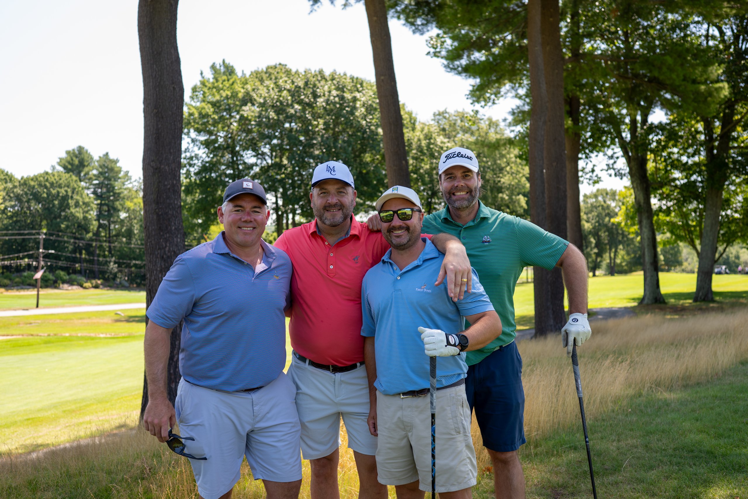Foursome of four men posing arm in arm on the tee beneath tall pines, the fairway behind them.