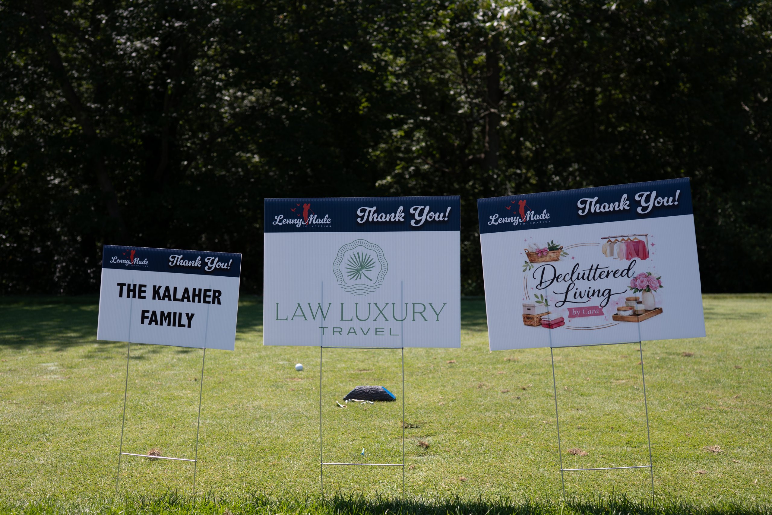 Three LennyMade Foundation sponsor signs lined up at a tee: The Kalaher Family, Law Luxury Travel and Decluttered Living by Cara.