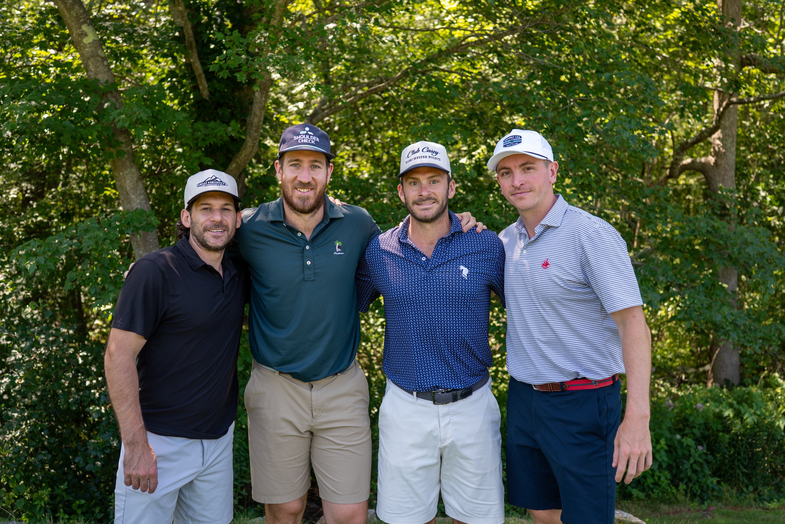 Foursome of four men in ballcaps posing arm in arm along the wooded edge of the course.