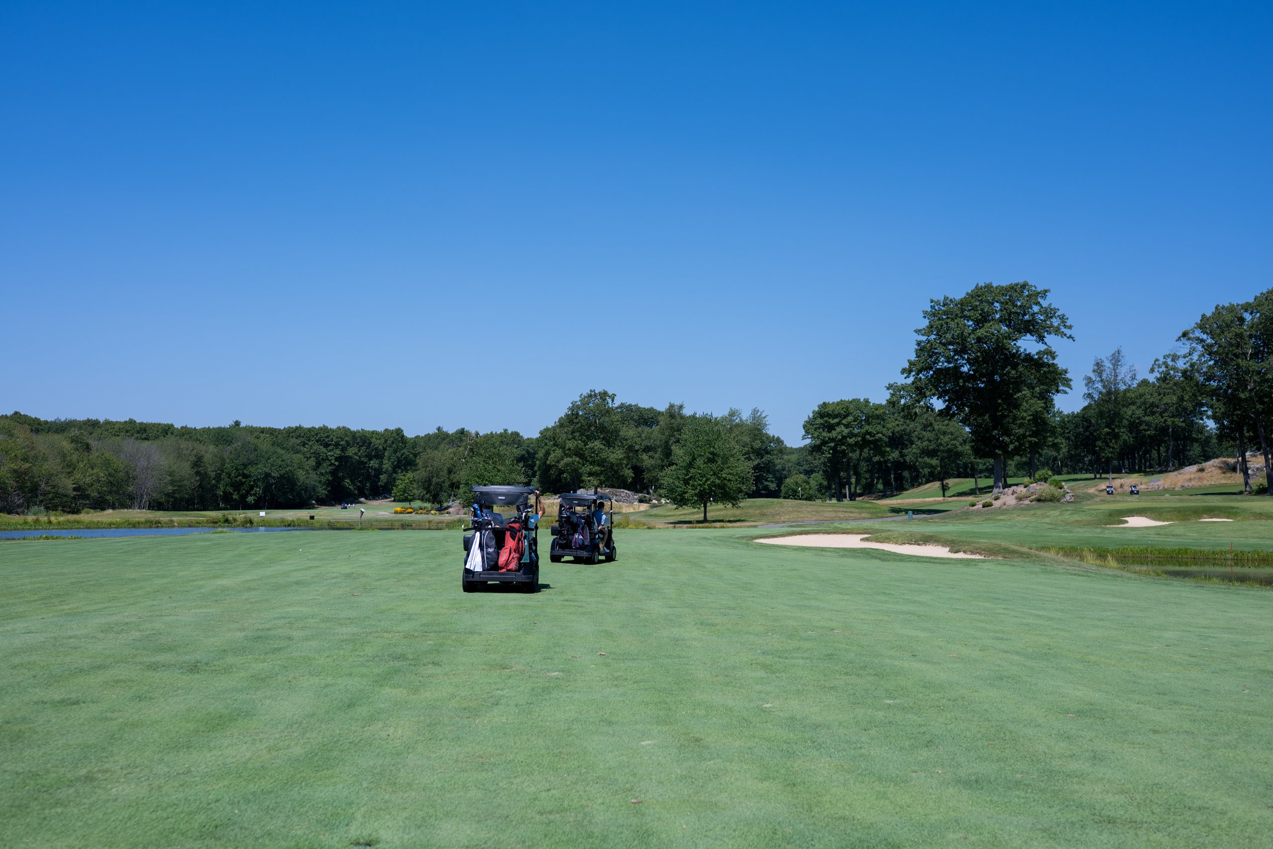 Two golf carts heading down a wide fairway toward the green, a bunker and pond off to the right.