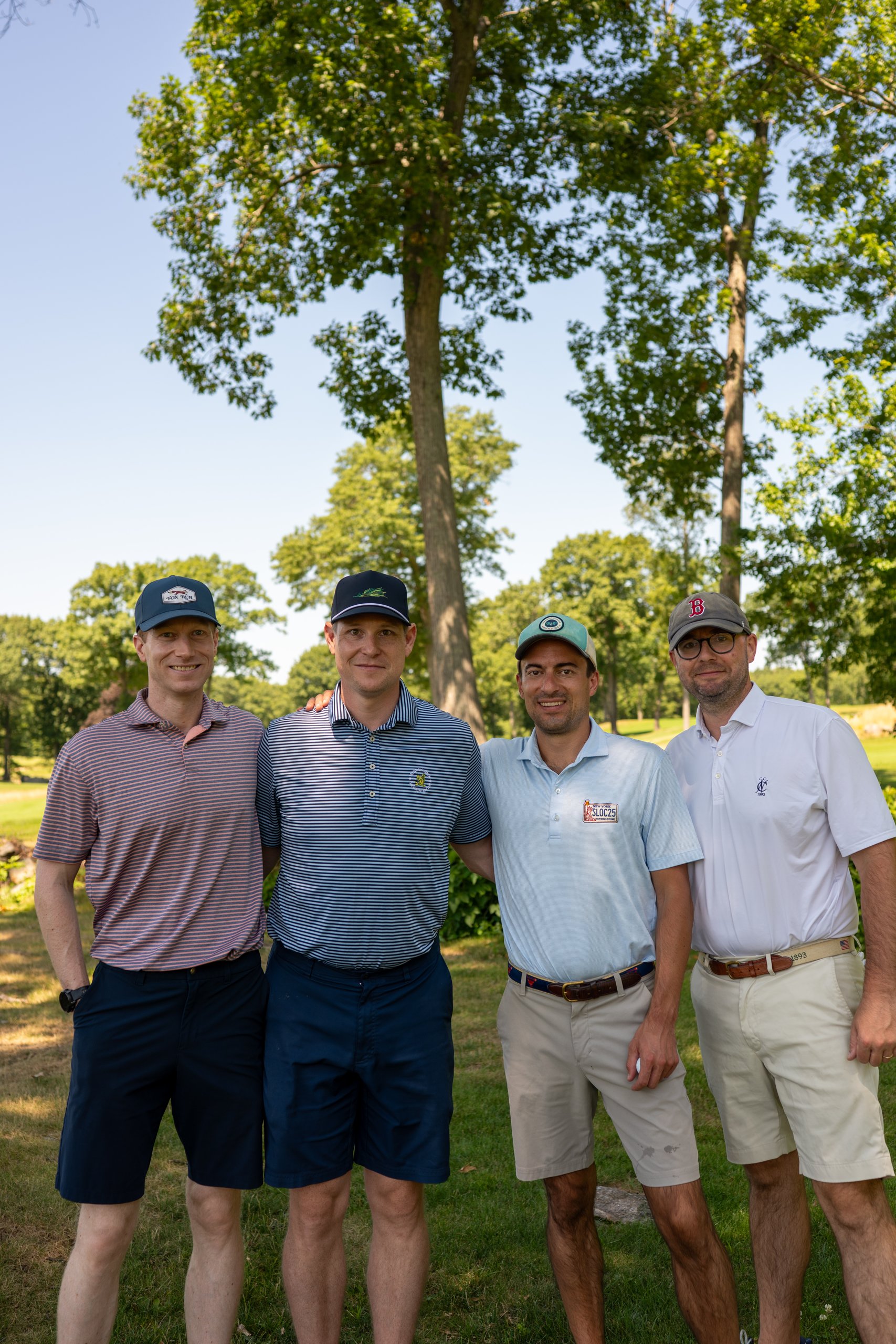 Foursome of four men posing arm in arm beneath a large tree, the fairway stretching behind them.