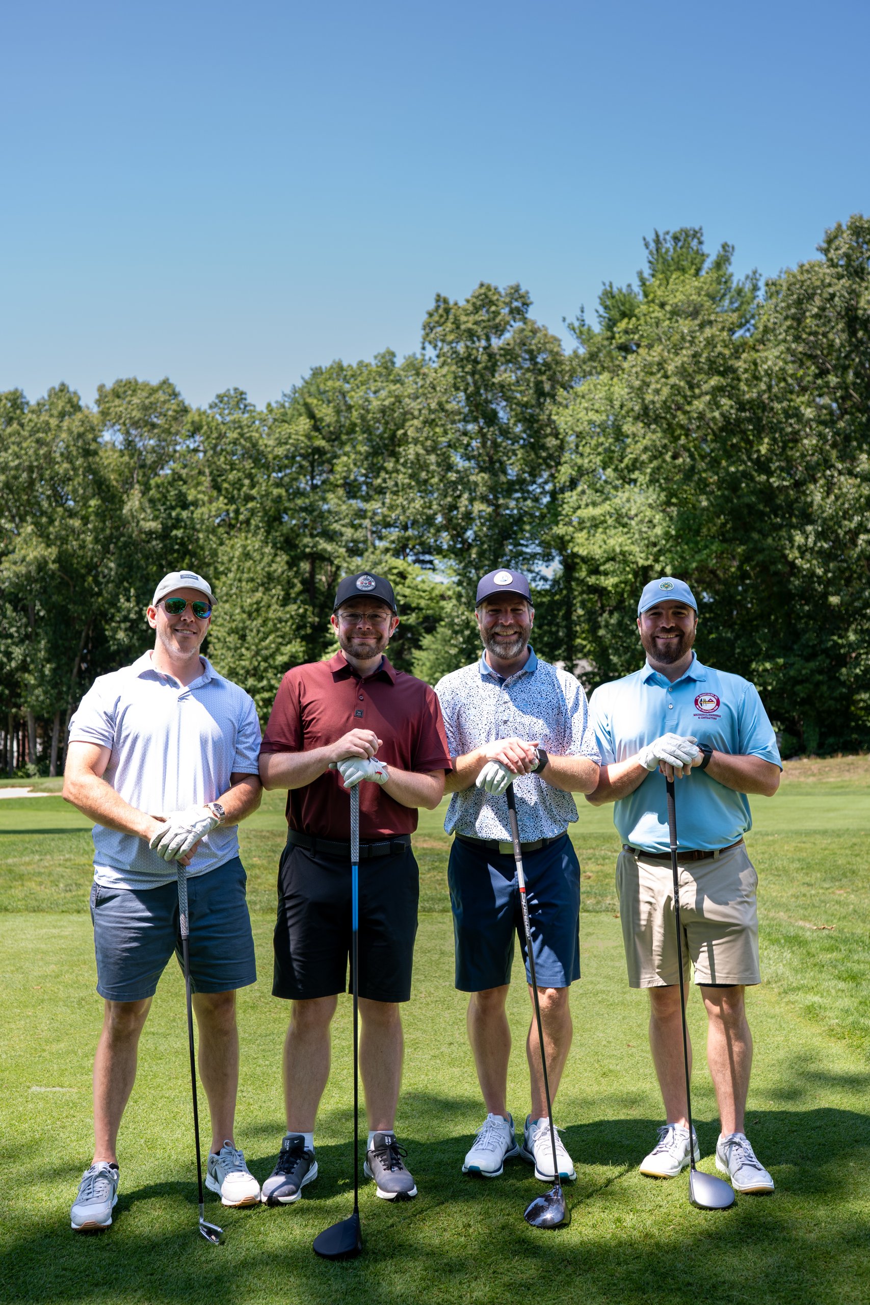 Foursome of four men posing with their drivers on the tee, tall trees filling the background.