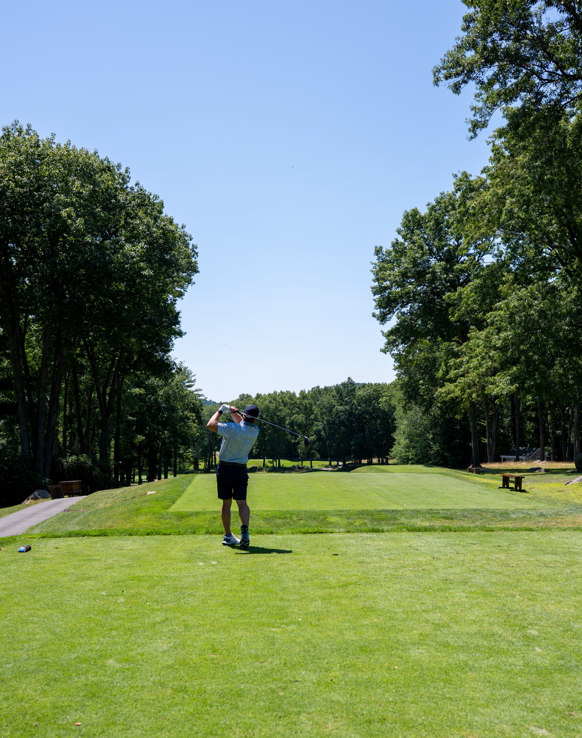 Golfer in a blue polo finishing his tee shot, looking down a long tree-lined fairway.