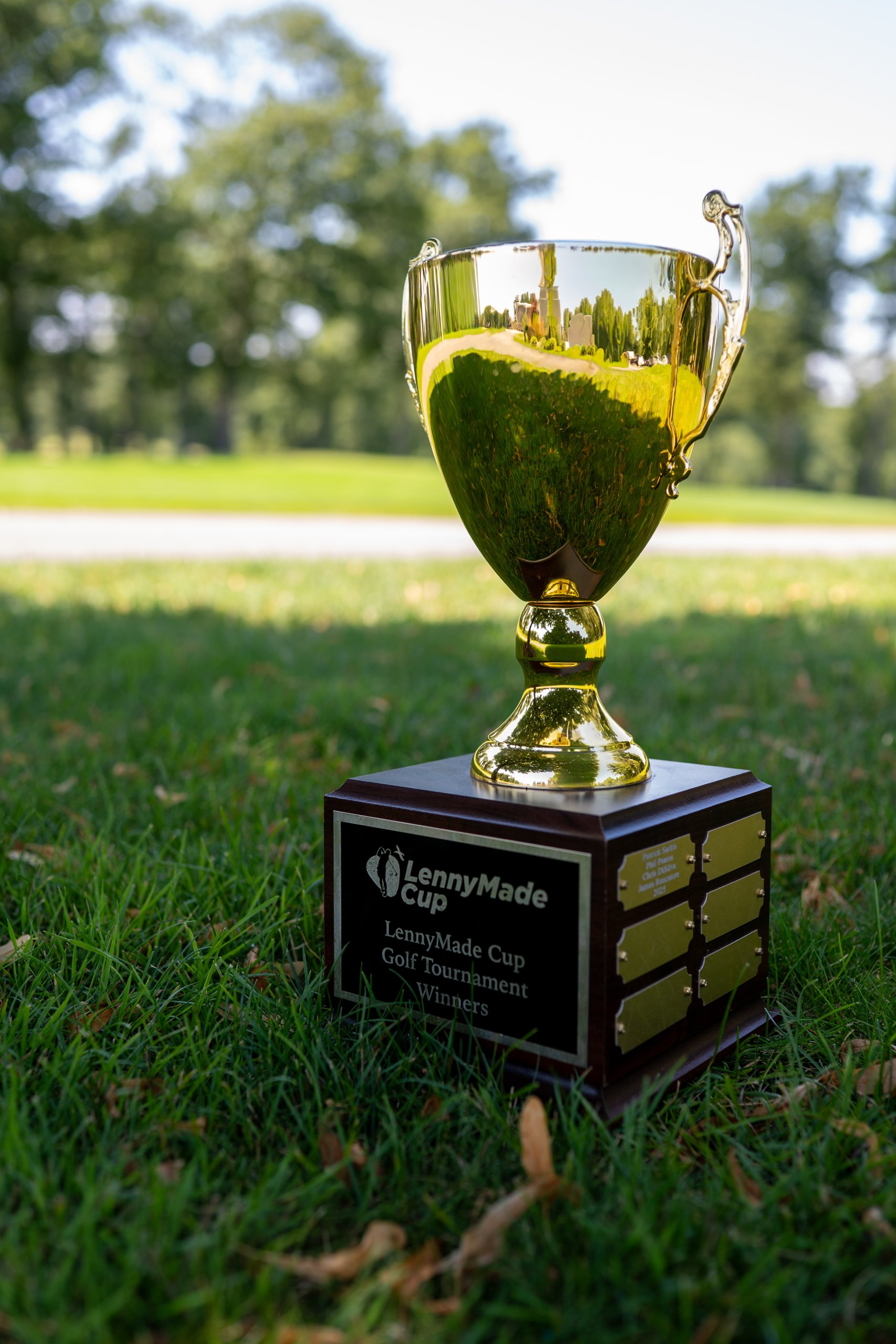 The gold LennyMade Cup perpetual trophy sitting in the grass, its base plaque reading LennyMade Cup Golf Tournament Winners.