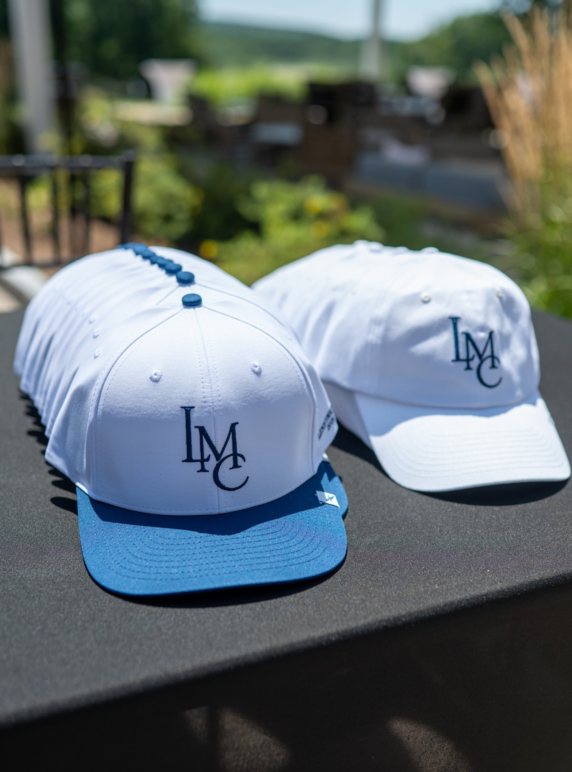 A stack of white LMC-monogram caps, one with a blue brim, displayed on the merchandise table.