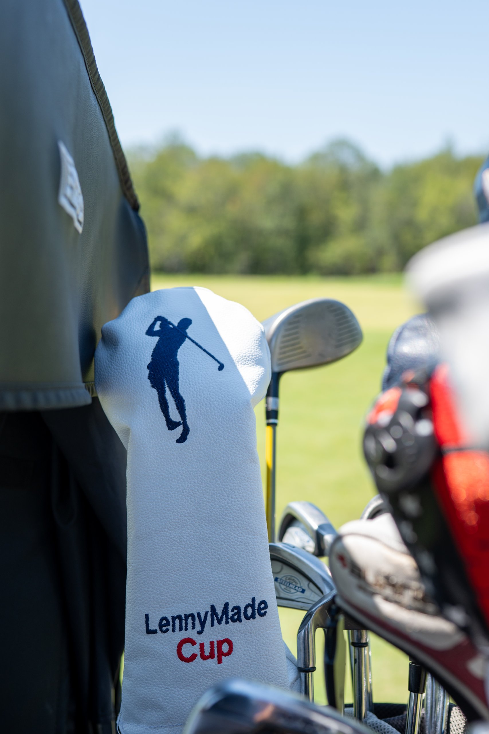 A white LennyMade Cup driver headcover standing up in a golf bag beside the clubs on a cart.