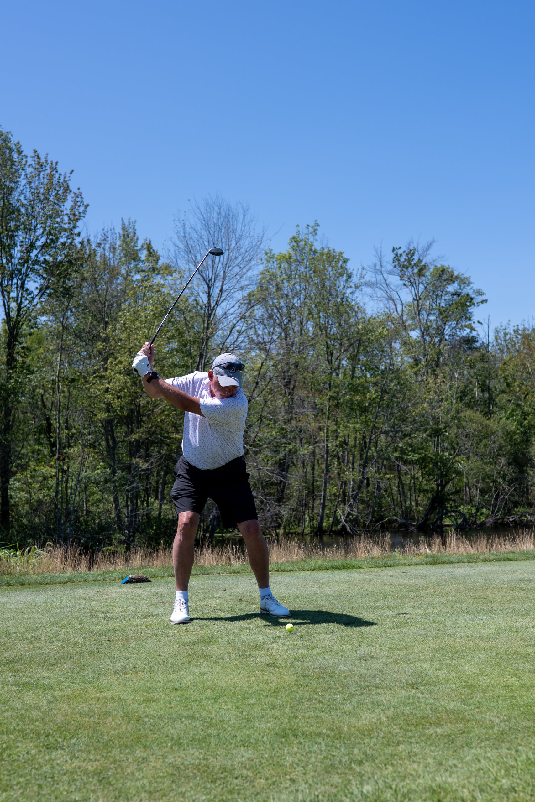 Golfer in a white polo and cap at the top of his backswing on the tee, a marshy pond and trees behind.
