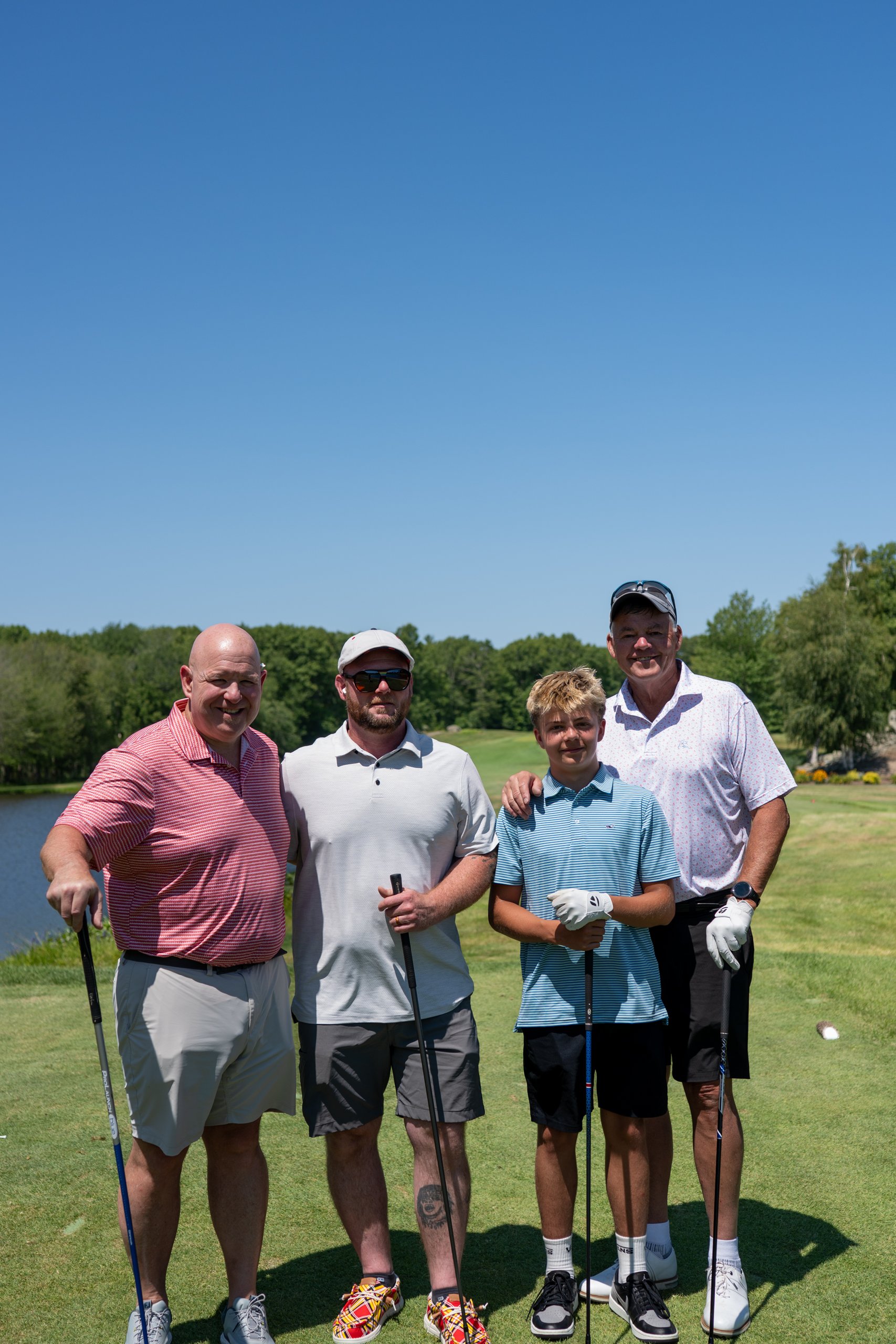 Foursome of three men and a boy posing with their clubs at the tee, a pond and treeline behind.