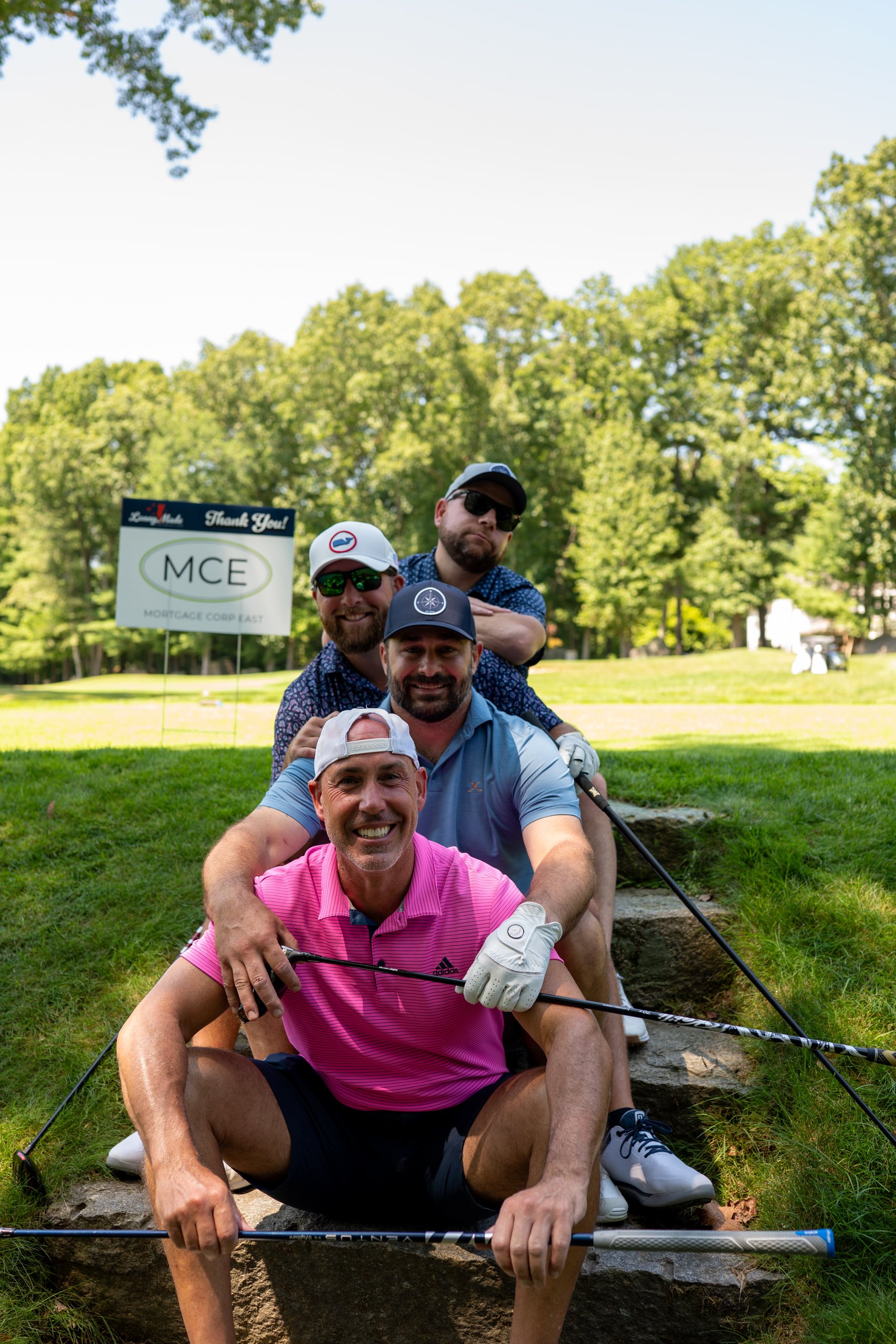 Four golfers stacked playfully on stone steps with their clubs, an MCE Mortgage Corp East sponsor sign behind.