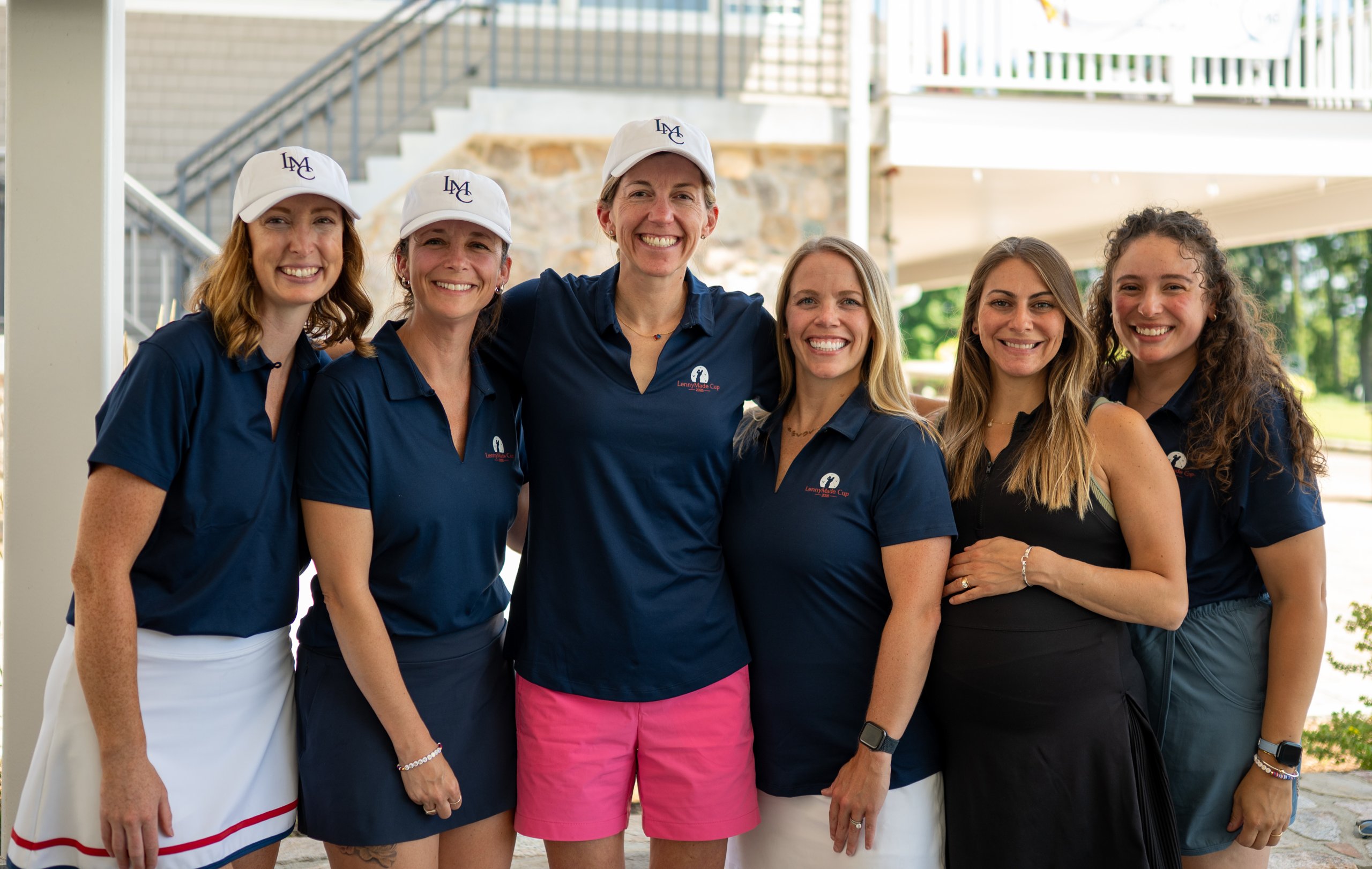 Six women in navy LennyMade Cup polos and LMC caps posing together on the clubhouse porch.