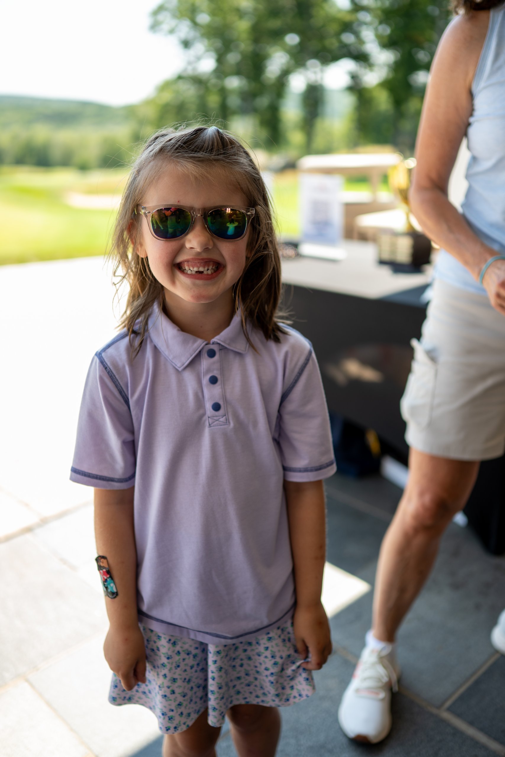 A young girl in a lavender polo and mirrored sunglasses grinning on the patio.
