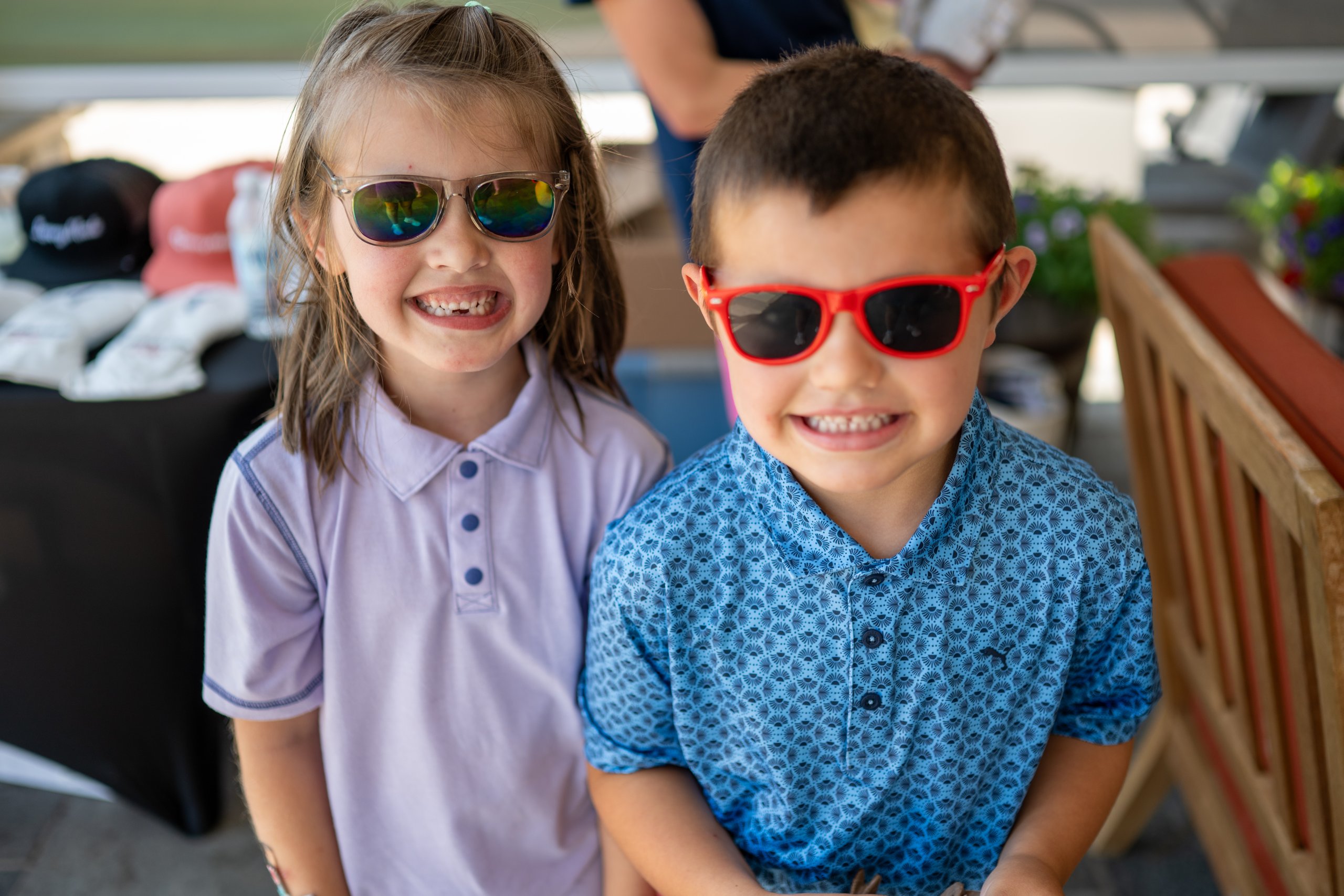 Two children in sunglasses smiling together on the patio, one in lavender and one in a blue polo.