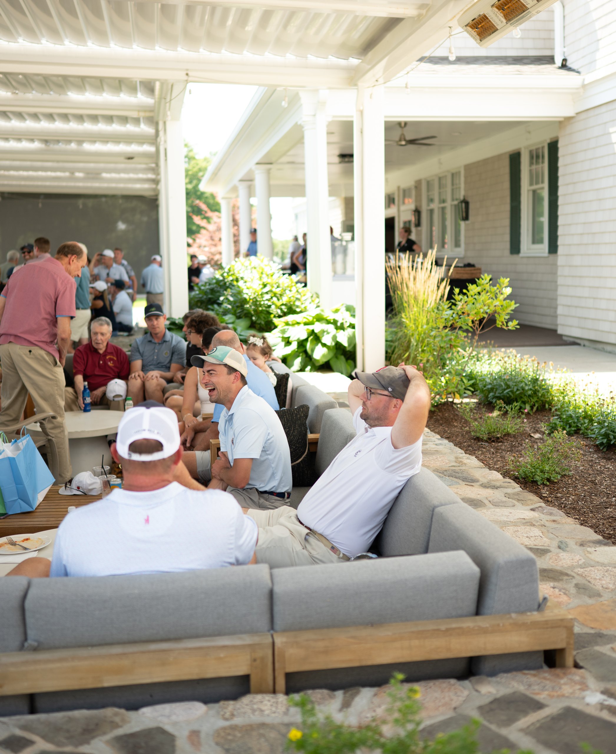 Guests relaxing and laughing on grey patio couches under the clubhouse pergola.