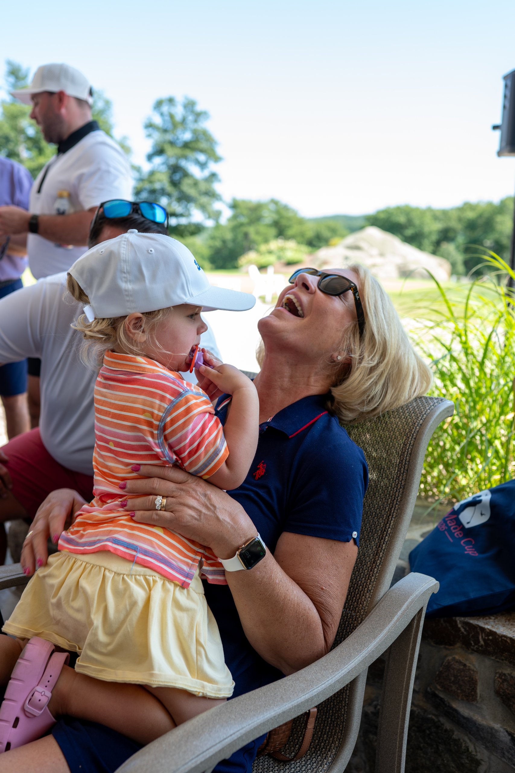A woman in a navy polo laughing with a toddler on her lap on the clubhouse patio.