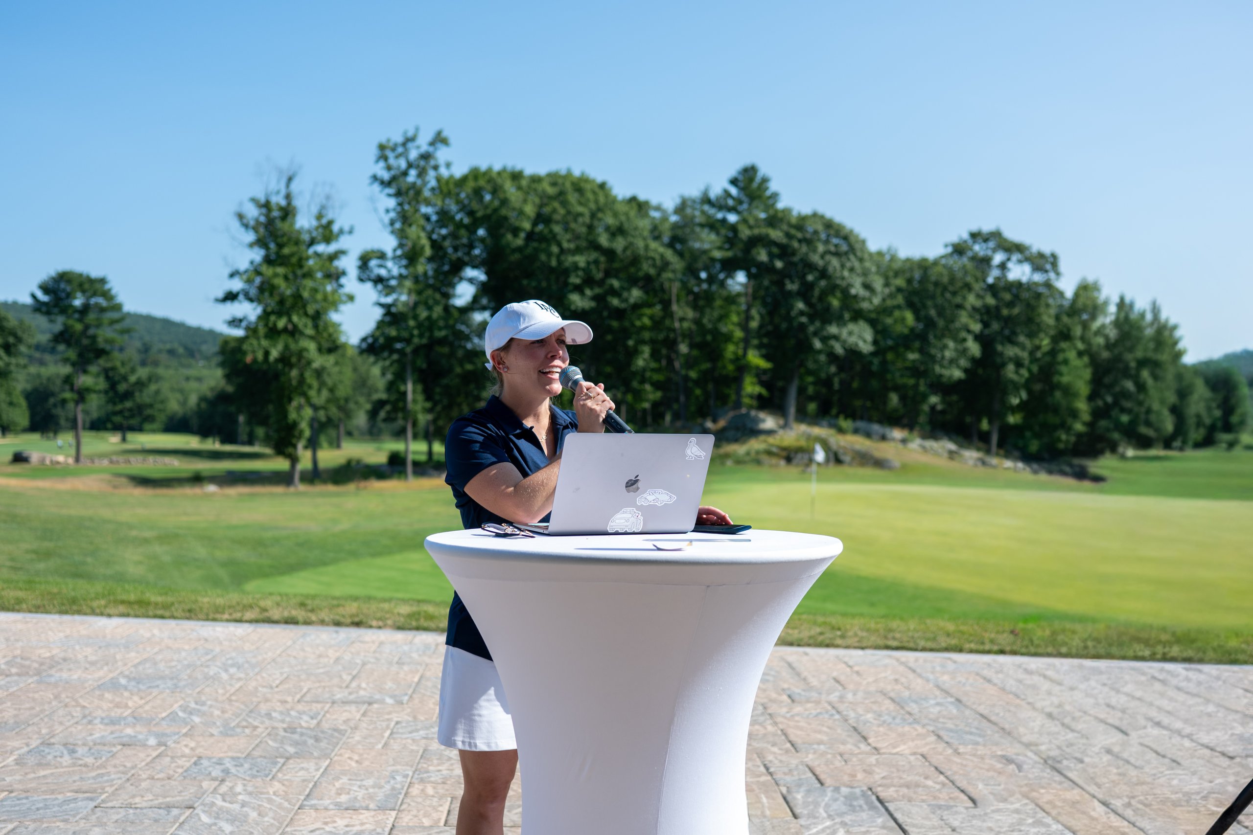 Event host in an LMC cap speaking into a microphone at a laptop podium, the course behind her.