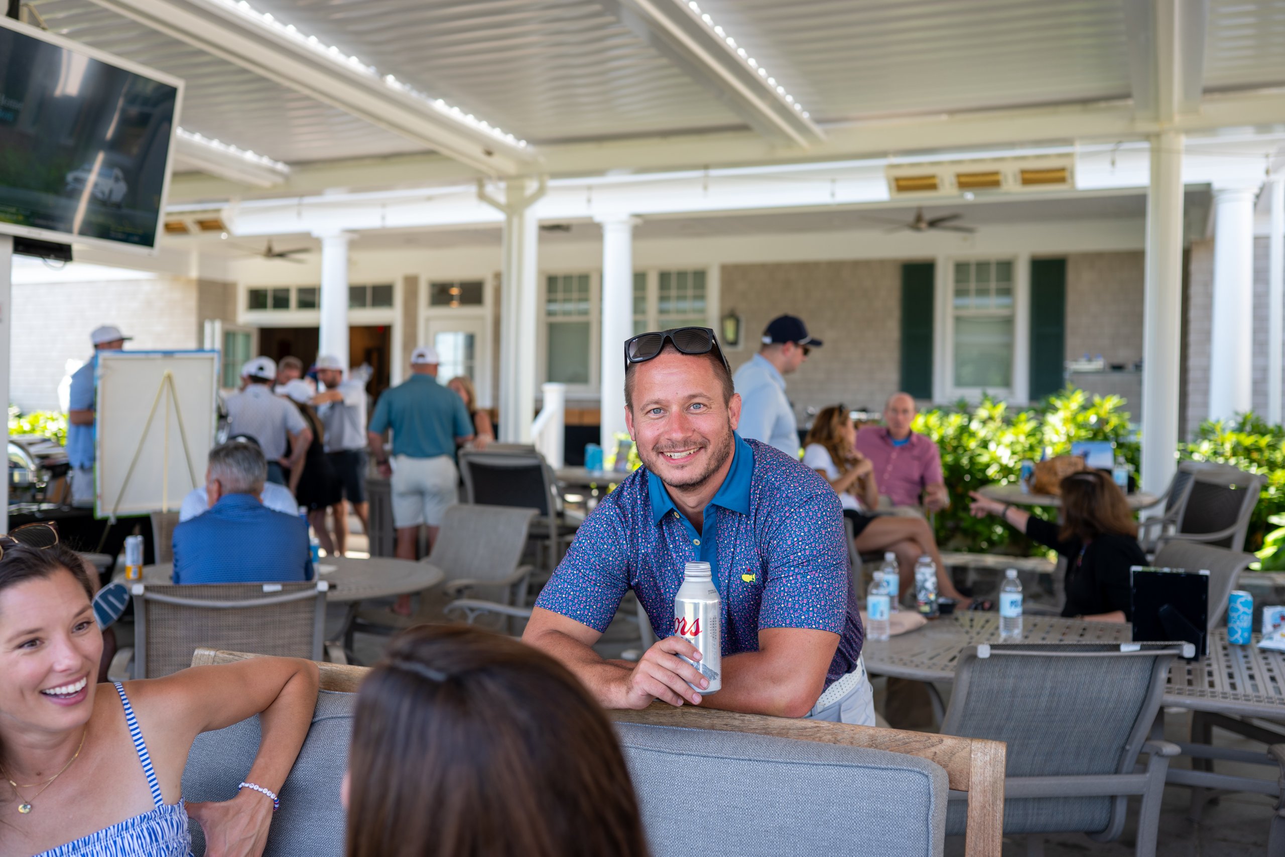 A guest in a patterned polo smiling and leaning on a patio chair with a drink, others mingling behind.