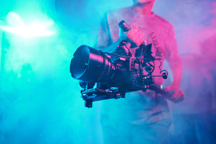 A camera operator holding a professional film camera rig on a smoke-filled set lit in blue and pink