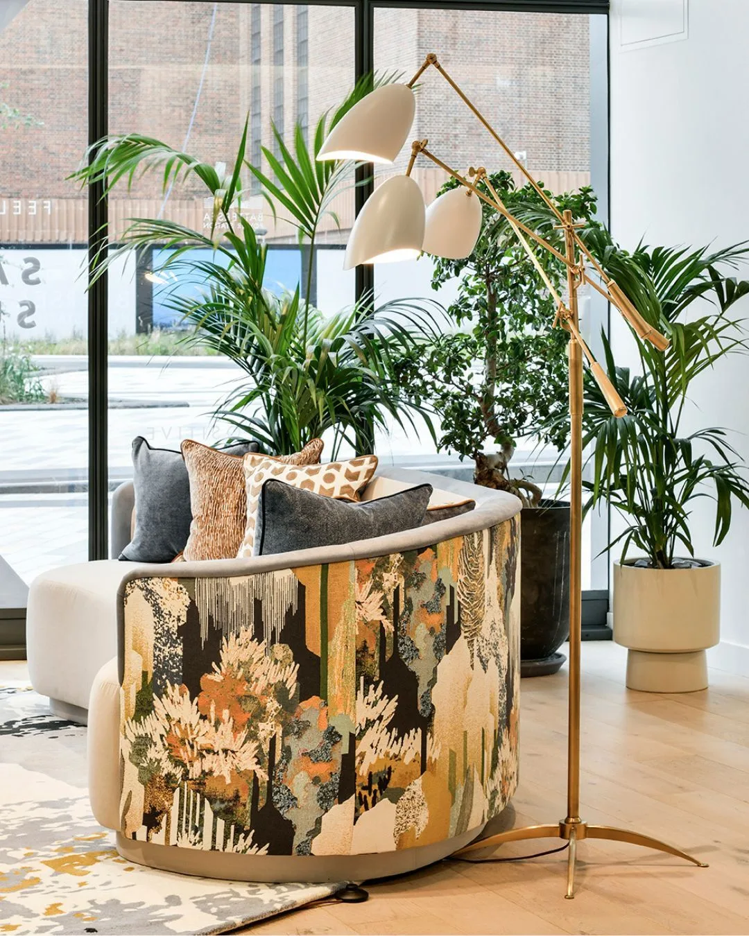 A bright interior corner featuring a curved modern sofa with an abstract nature-patterned back, a brass three-armed floor lamp, and large potted plants.