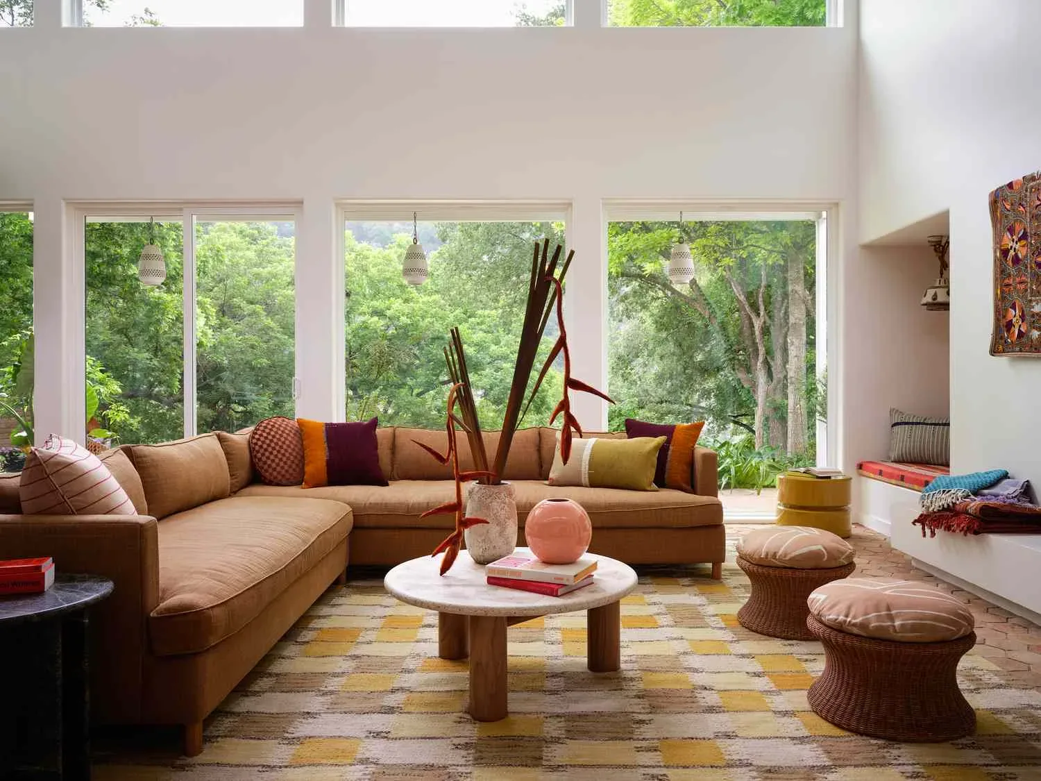 Spacious living room with a caramel sectional sofa, earth-tone accent pillows, round wooden coffee table, patterned rug, and floor-to-ceiling windows overlooking lush greenery, designed by Decozie designer Nicole W.