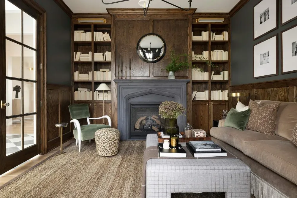 Sophisticated living room with dark wood paneling, built-in bookshelves, a black fireplace, neutral sofa with green accent pillows, and a cozy reading chair, designed by Decozie designer Austin T.