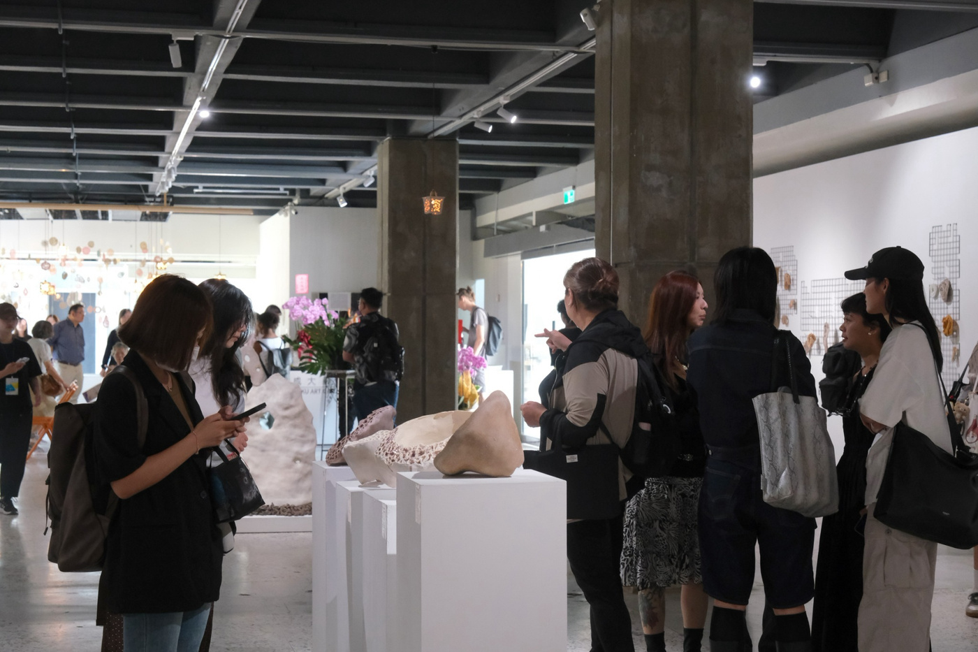 一個擁擠的藝廊開幕式，許多訪客正在檢視展示在白色展台和牆面上的藝術作品。巨大的混凝土柱支撐著天花板，紫色花卉裝飾著空間。背景可見懸掛的雕塑作品。