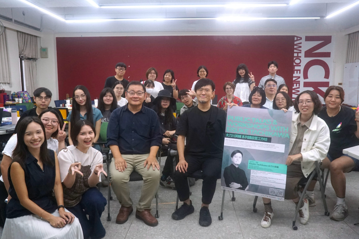 A large group of approximately 25 young people posing together in a bright classroom with a maroon accent wall. They are holding a research poster with green and teal text and graphics. The room has modern LED lighting and classroom furniture visible in the background.