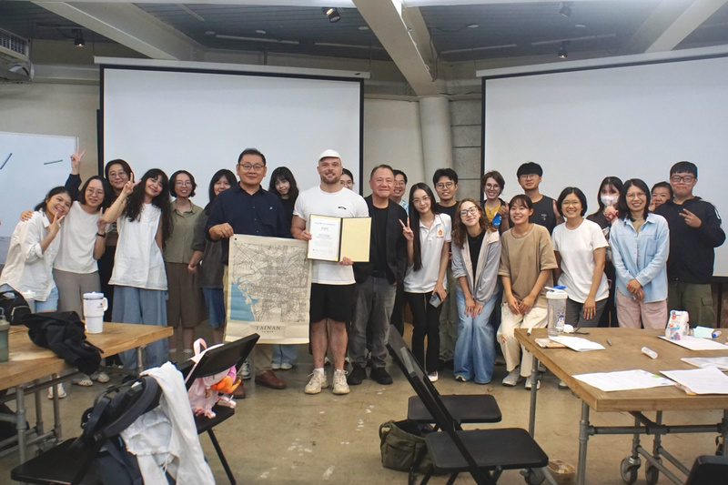 約25名學生與講師在教室內合影，中間有兩人手持一幅裝裱好的證書和一張台南的老地圖。背景中可見投影螢幕和木製桌子。